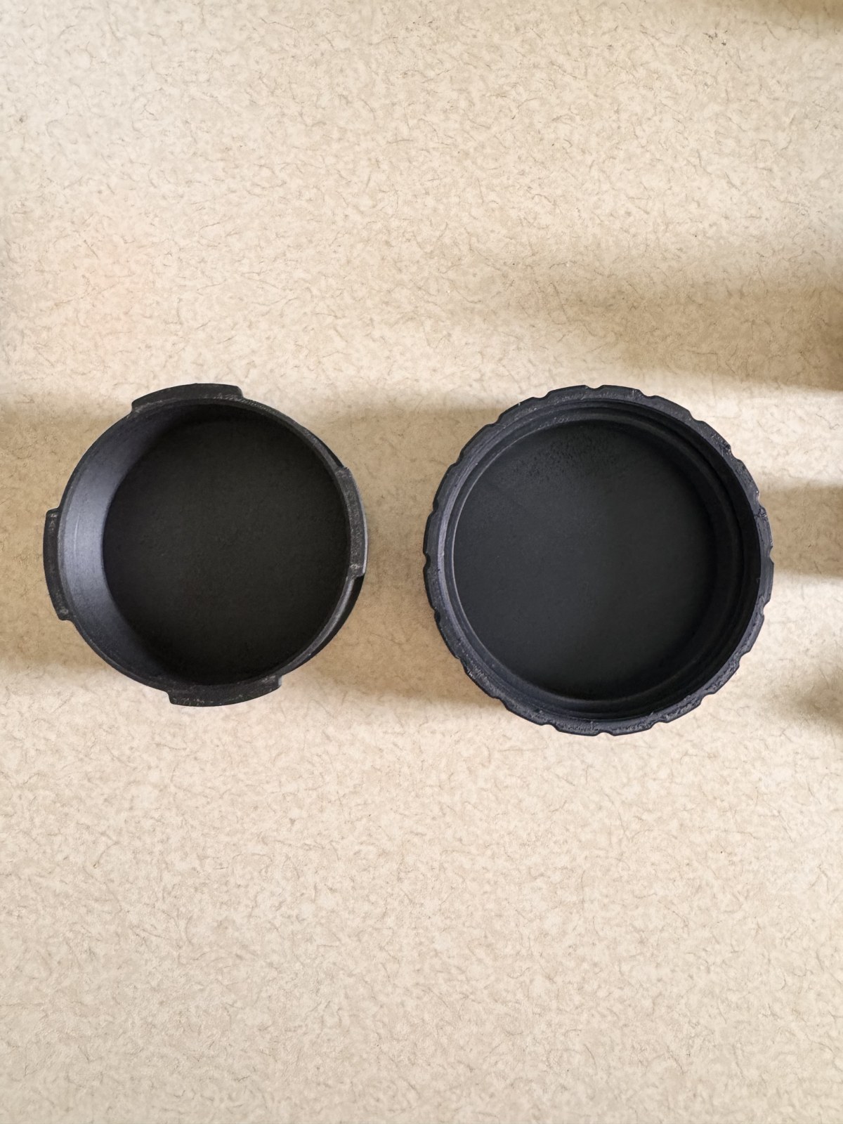 The two finished halves of the AeroPress filter container sitting on a white surface, the shorter ridged bottom on the left and the smooth-walled lid on the right, both printed in matte black PLA