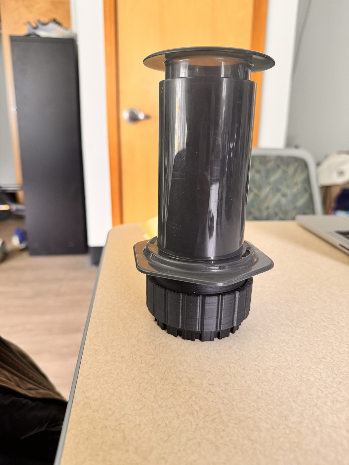 The assembled AeroPress unit standing on a desk with the printed filter container clamped onto the plunger end, the container reading as a base that the rest of the brewer extends out of, paper filters stored inside