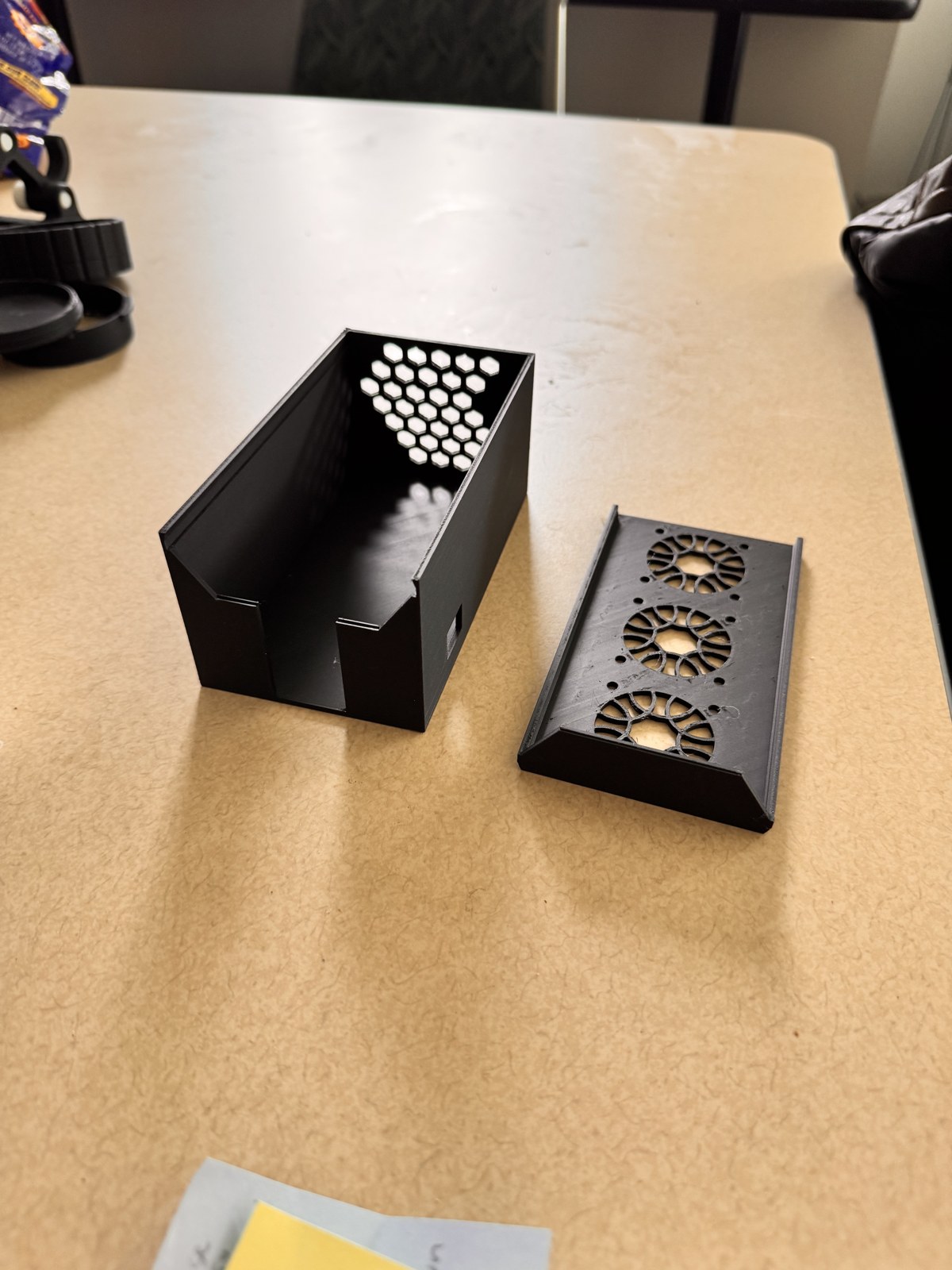 The cooling box shell on the left, hollow with a hex pattern of vents on the back wall, and the separate top plate on the right with three round fan cutouts and a grid of bolt holes, both printed in matte black PLA