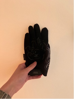 A black tactical-style glove with subtle gray graphics on the back held up against a warm beige wall, the wearer's bare forearm extending out of frame at the bottom of the image