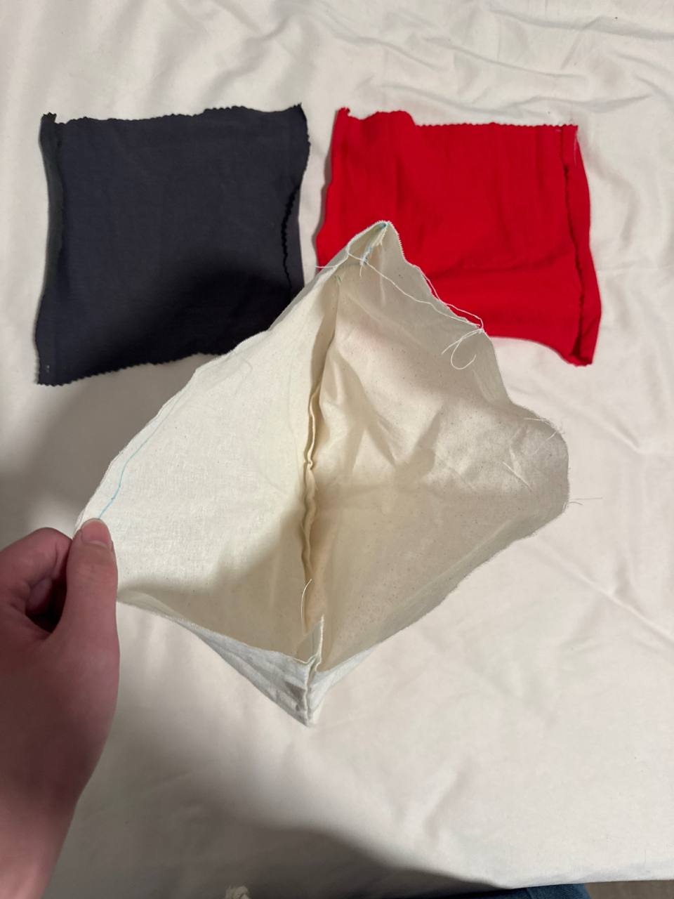 The finished muslin prototype 1 held open by hand to show the interior cavity, with the unused 7 by 8 inch dark grey and red pocket pieces visible in the background