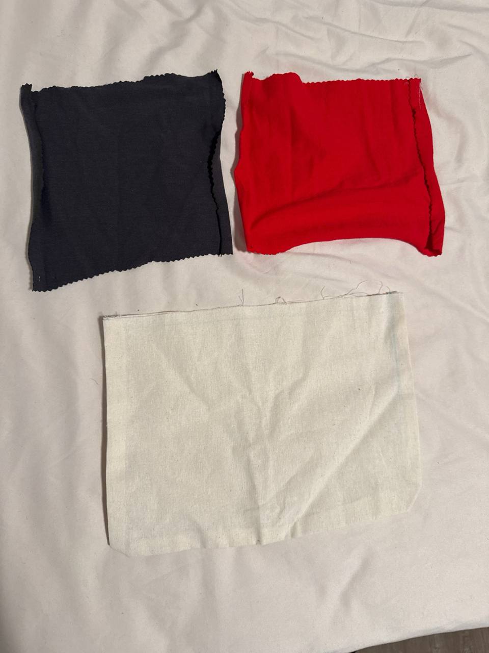 The unused interior pocket pieces laid flat on a white sheet, cut but never sewn into the sleeve: a dark grey 7 by 8 inch t-shirt rectangle and a red 7 by 8 inch t-shirt rectangle in the top row, with an off-white muslin offcut below them
