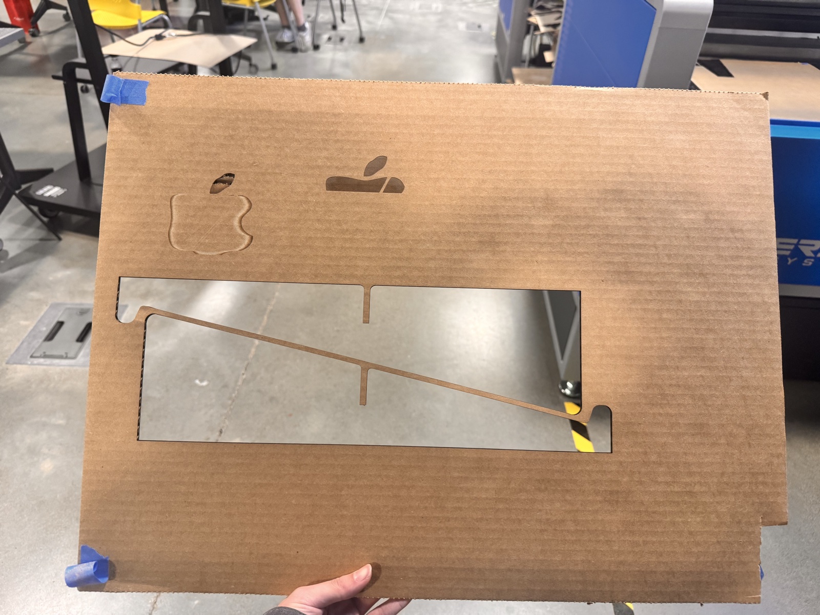 The laser cutter bed with the finished flute board cut sheet still in place, showing one large rectangular cutout where the two side panels and surface details have been lifted out, with the remaining material showing the engraved wax apple silhouette and a smaller cut-through version of the same symbol near the top corner