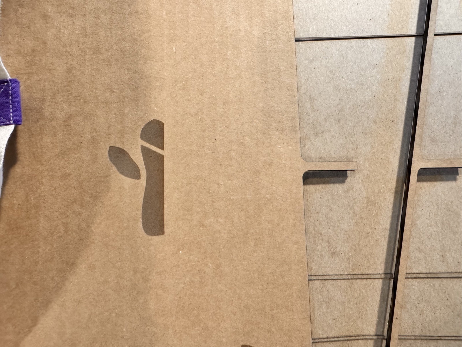 Close-up of a flute board panel still on the laser bed showing a small wax apple silhouette that has only been scored along its outline rather than cut through, with the surrounding panel showing finished slot cuts, evidence of the premature shutdown that left this single feature incomplete