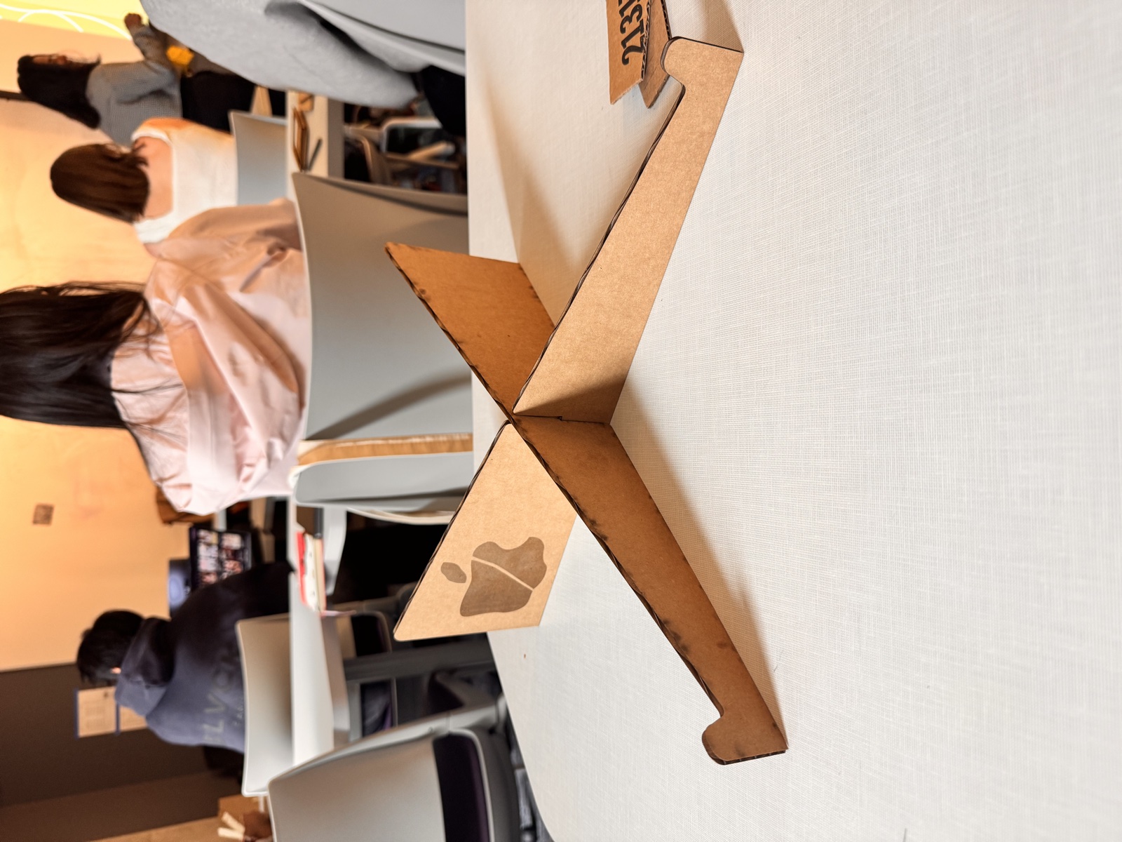 The finished laptop stand assembled, the two flute board panels interlocking at ninety degrees to form an X-shape standing upright on a white studio table, with the engraved wax apple symbol visible on the front face and other students working in the background