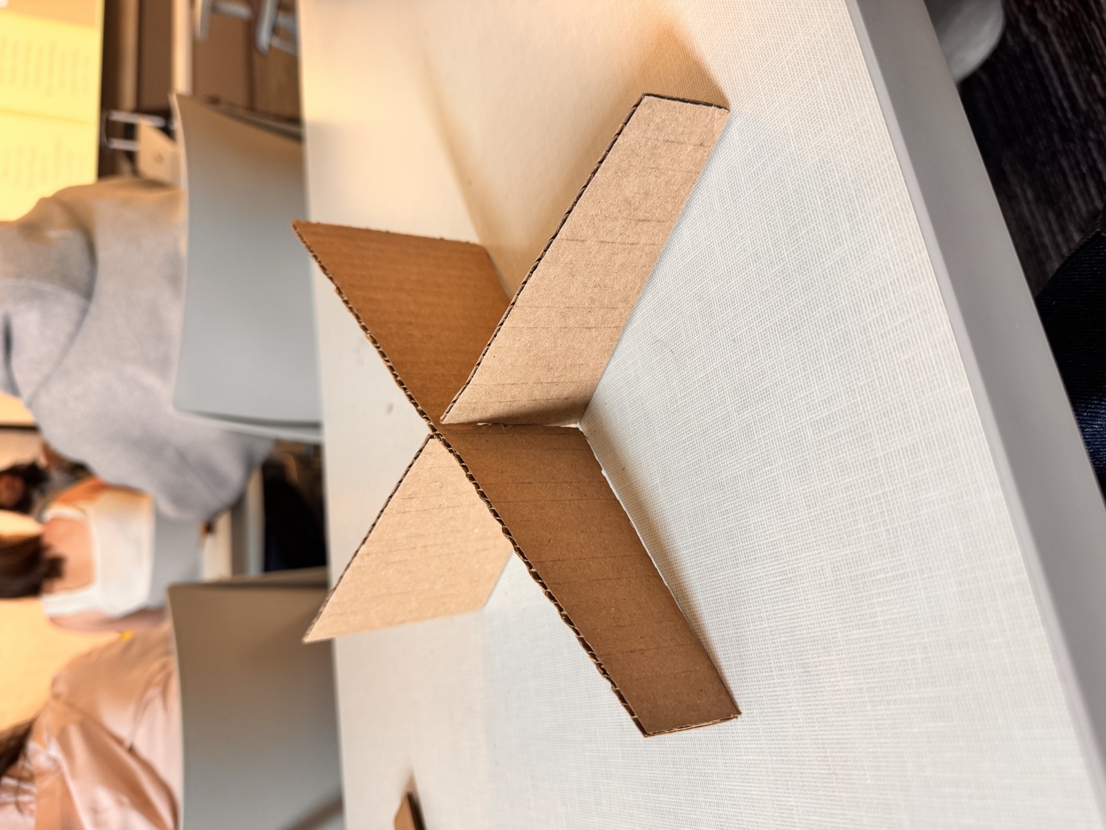 The two cardboard panels slotted together at ninety degrees forming an X-shaped laptop stand sitting upright on a white desk in the HCDE studio, with the corrugated edges visible along the top