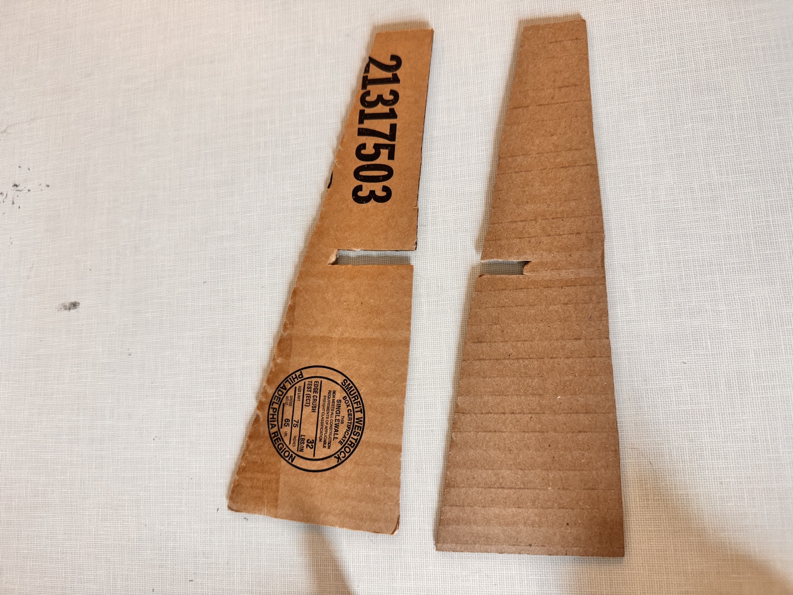 Two wedge-shaped panels cut by hand from a recycled Amazon shipping box, laid flat on a white surface, each with a center slot cut into one long edge, the panel on the left still showing a printed shipping label and barcode, and the panel on the right showing the corrugated grain of the cardboard