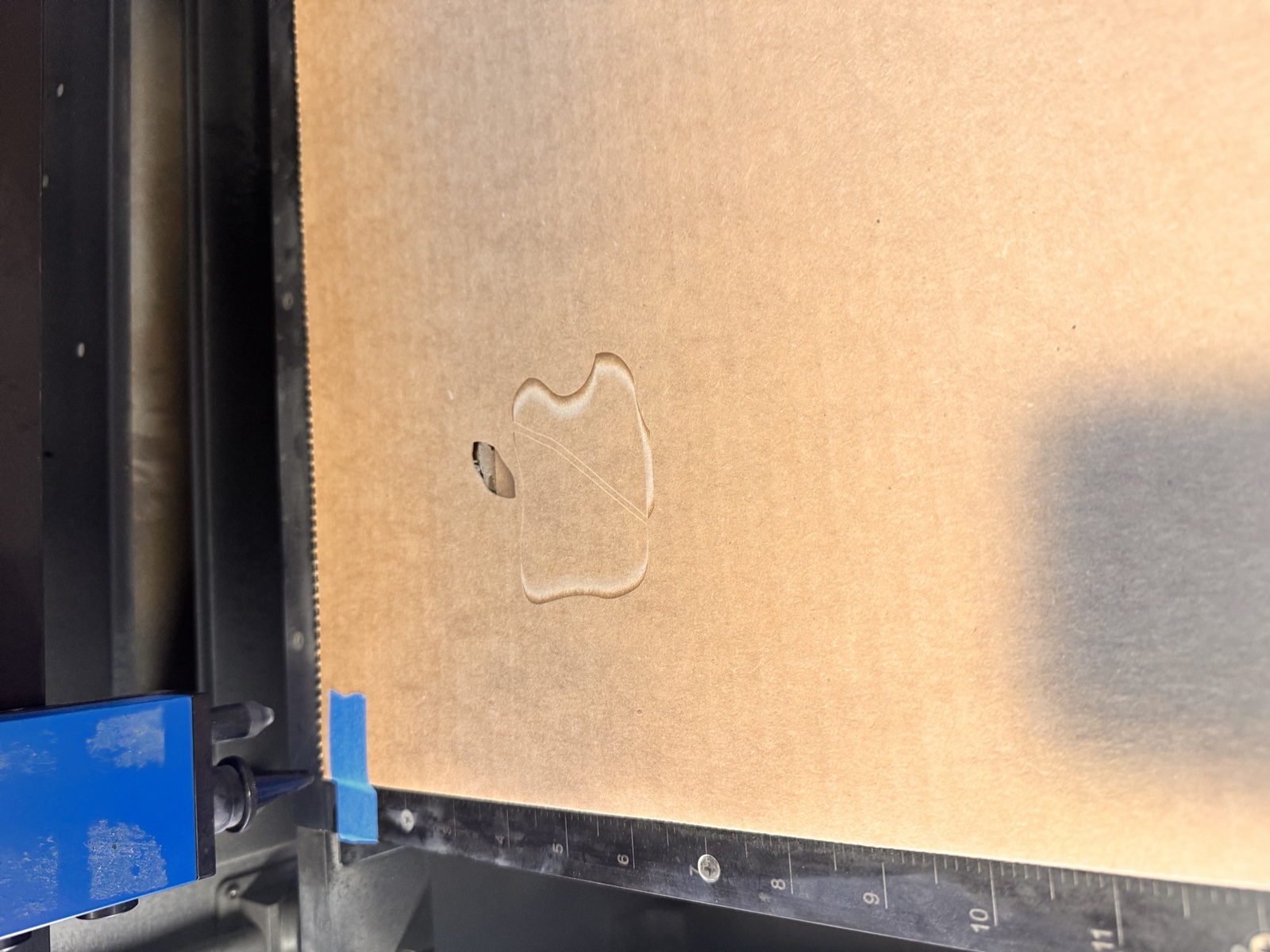 Close-up of a flute board surface inside the laser bed showing the wax apple engraving where the perimeter has been overburned into a deep brown halo and the leaf detail has charred almost black, the result of pushing the engrave pass to 35 percent power during the calibration walk-up