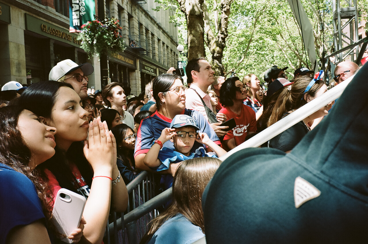 A crowd gathered along a tree-lined street, with spectators behind a barrier.