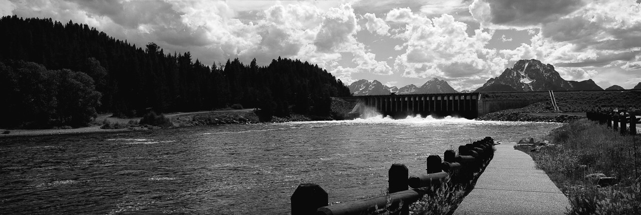 A black-and-white lakeside panorama with wooden posts leading toward distant mountains.