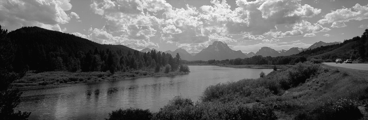 A black-and-white panoramic landscape with water, trees and distant mountains.