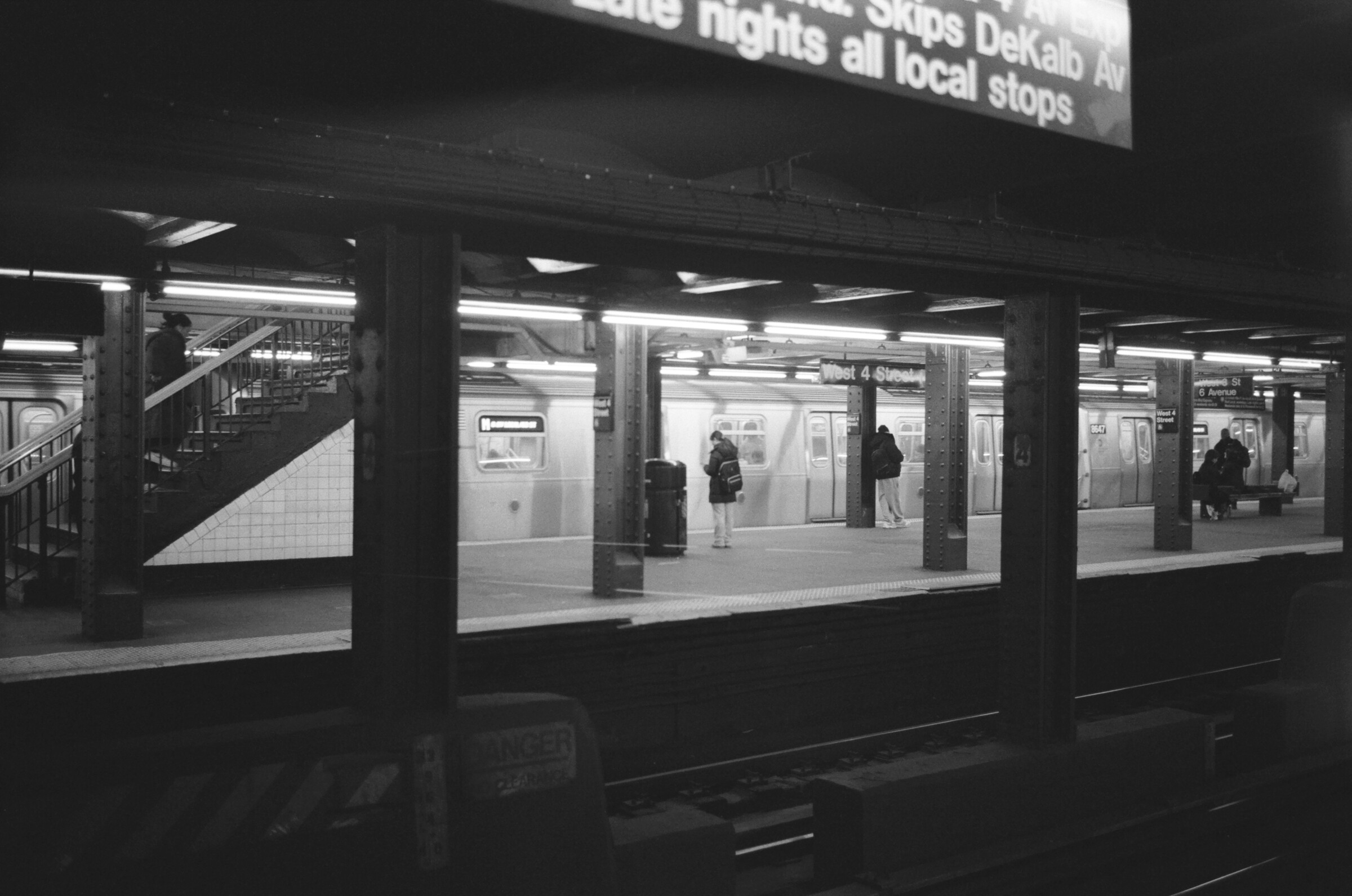 Black-and-white view across subway tracks toward a platform beneath overhead signs.