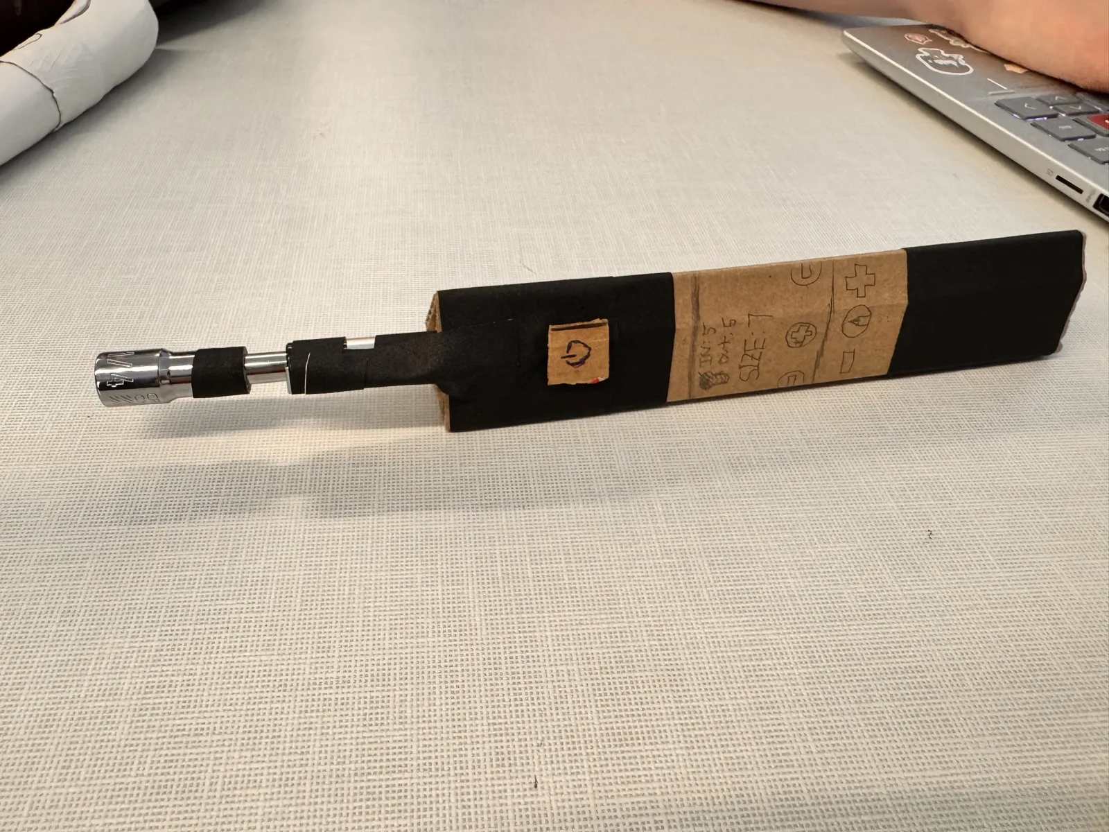 Angled view of the cardboard prototype on a desk showing the pen shaft extending from the handle with the socket chuck at the tip
